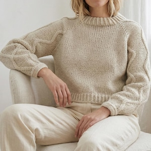 Op de afbeelding: Een beige, grofgebreide trui met een ronde hals en raglanmouwen. De mouwen hebben een kabelgebreid patroon. De trui wordt gecombineerd met een beige broek. Het model zit op een stoel.