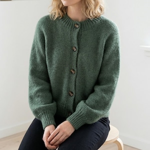 May include: A green knit cardigan with a round neckline and button-up closure. The long-sleeved sweater has ribbed cuffs and a relaxed fit. The buttons are a brown color. The cardigan is made of a soft, textured yarn.