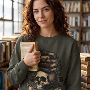 Puede incluir: Sudadera verde oliva con un diseño de calavera, libros y rosas con el texto "READ IN PEACE". Una persona lleva la sudadera y sostiene un libro. Se ven estanterías llenas de libros en el fondo.