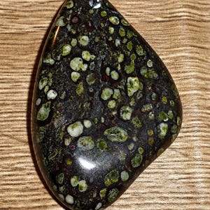 May include: A polished, teardrop-shaped pendant featuring a dark, nearly black base stone speckled with numerous rounded inclusions in varying shades of light green, olive, and white, attached to a silver-toned metal bail at the top.