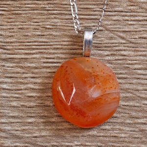 May include: A round, translucent orange carnelian pendant with visible reddish-brown speckles and natural internal veining. The stone is secured by a silver-toned metal bail and hangs from a matching silver-toned metal figaro chain necklace.