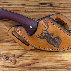 May include: A fixed-blade knife with a dark reddish-brown wooden handle and two brass rivets, housed in a tan leather sheath featuring a laser-engraved deer head design, secured with copper-colored metal rivets and featuring integrated belt loops.