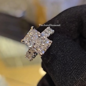 Puede incluir: Un deslumbrante anillo de compromiso de diamantes con una gran piedra central de corte cuadrado y diamantes de acento más pequeños. El engaste es probablemente de platino u oro blanco, con un acabado pulido. The Aura Diamond es visible.
