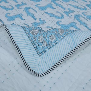 Pode incluir: Um edredom branco com um padrão repetido de crocodilos azul claro. O edredom tem uma borda azul clara com um desenho floral e uma borda listrada em preto e branco. O edredom está dobrado, mostrando a parte inferior.