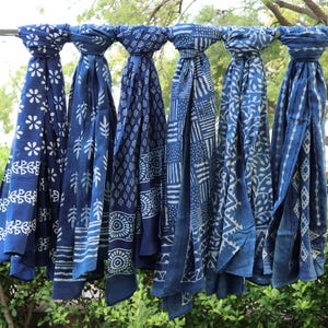 Puede incluir: Seis bufandas de tela rectangulares colgadas una al lado de la otra en una barra de metal. Cada bufanda presenta un patrón blanco distinto impreso sobre un fondo azul profundo, incluyendo motivos florales, geométricos, rayas y hojas.