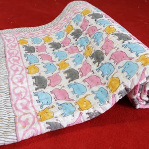Pode incluir: Um cobertor acolchoado enrolado com fundo branco e um padrão repetido de elefantes coloridos em rosa, azul, amarelo e cinza. A borda apresenta um design floral rosa e um padrão listrado.