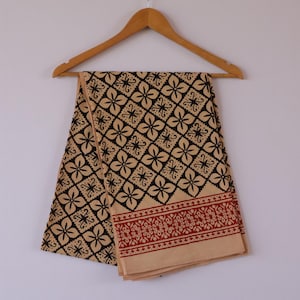 Op de afbeelding: Een beige en zwart patroontextiel hangt aan een houten kleerhanger. De stof heeft een herhalend bloemenpatroon in zwart en een decoratieve rode rand. Het textiel is waarschijnlijk een sjaal of omslagdoek.