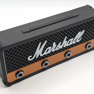 May include: Black and copper-colored miniature amplifier with the word "Marshall" in white script across the front. The amplifier has a handle on top and three black knobs. The front has a speaker grill design.