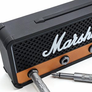 May include: A black and copper-colored miniature amplifier with the word "Marshall" in white script. Two silver-colored jack plugs are partially inserted into the amplifier. The amplifier has a speaker grill design.