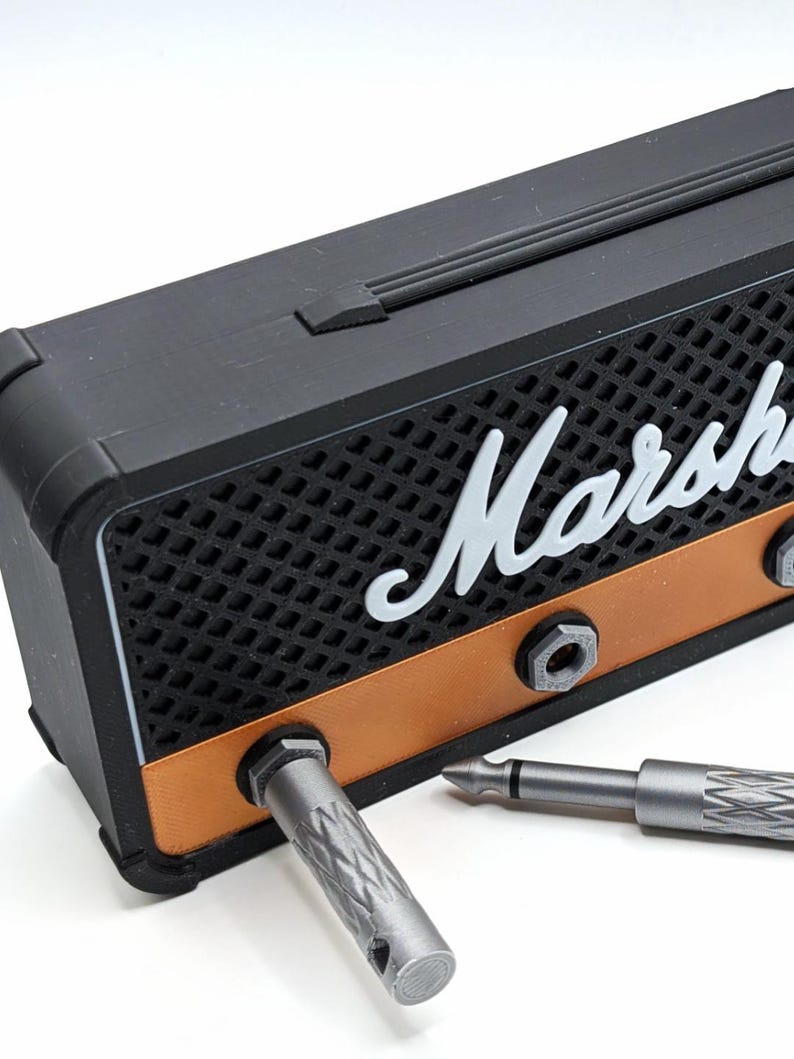May include: A black and copper-colored miniature amplifier with the word "Marshall" in white script. Two silver-colored jack plugs are partially inserted into the amplifier. The amplifier has a speaker grill design.