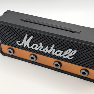 May include: A black and orange miniature amplifier with the word "Marshall" in white script. The front features a black mesh grill, orange accents, and silver-colored knobs. A black handle is on top.
