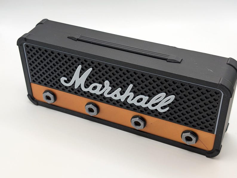 May include: A black and orange miniature amplifier with the word "Marshall" in white script. The front features a black mesh grill, orange accents, and silver-colored knobs. A black handle is on top.