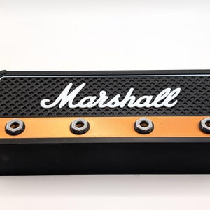 May include: A black and orange rectangular box with the white word "Marshall" across the top. The top has a black textured surface. There are three silver-colored, hexagonal nuts on the front. The box has a black border.