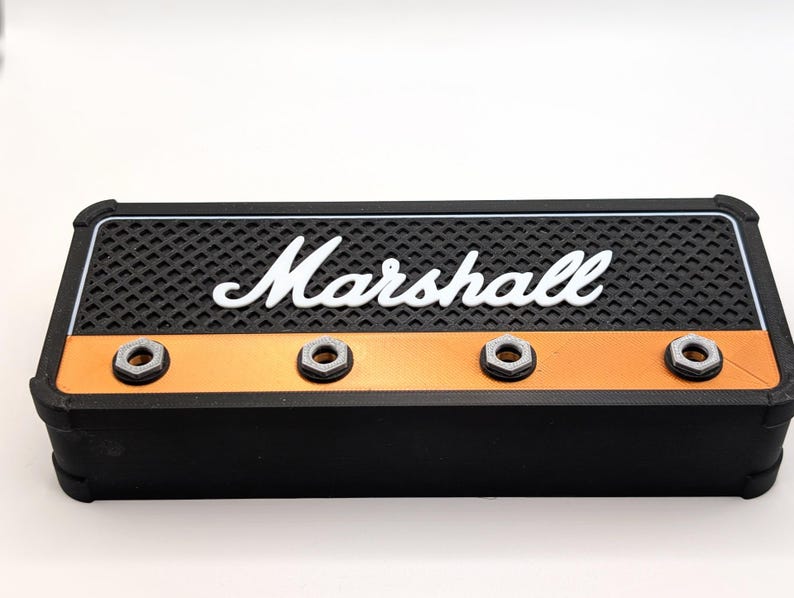 May include: A black and orange rectangular box with the white word "Marshall" across the top. The top has a black textured surface. There are three silver-colored, hexagonal nuts on the front. The box has a black border.