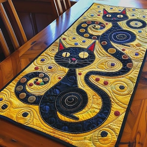 May include: A rectangular fabric table runner featuring two stylised black cats with large yellow eyes and red-lined ears. The background is a vibrant yellow with swirling patterns, circular motifs, and small colourful accents throughout the quilted design.