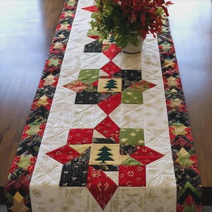 May include: A quilted table runner featuring a patchwork design with red, green, black, and white fabric squares. The center panels display appliqued dark green pine tree motifs on cream backgrounds, surrounded by star-shaped geometric patterns.