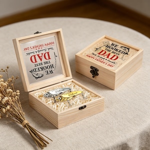 May include: Two light-colored wooden boxes with metal latches. One box is open, revealing two metallic fishing lures inside on wood shavings. The inside of the lid features the text: WE HOOKED THE BEST DAD, HAPPY FATHER'S DAY, and names.