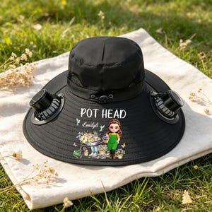 May include: A black sun hat featuring two small built-in fans on the brim. The front displays the text POT HEAD above a cartoon illustration of a person gardening with flowers, a wheelbarrow, and the name Emilyli printed in white script.