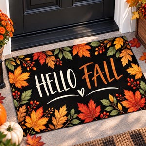 May include: A rectangular black doormat featuring the text HELLO FALL in white and orange lettering. The design is bordered by colorful autumn maple leaves in shades of red, orange, and yellow, along with small red berries on thin green stems.