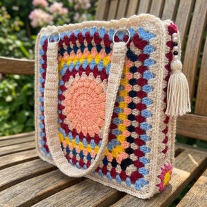 May include: A square, crocheted tote bag with a colorful, circular pattern in the center. The bag features a peach-colored center, surrounded by rings of yellow, blue, and burgundy. It has cream-colored straps and a tassel with beads.