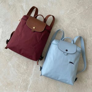 May include: Two nylon backpacks. One is burgundy with a brown leather flap and straps. The other is light blue with a matching flap and straps. Both have a top handle and adjustable shoulder straps.
