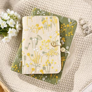 May include: Two embroidered journals with floral designs. The top journal is beige with yellow and white flowers, secured with a wooden button closure. The bottom journal is olive green with yellow and white floral embroidery. Both are on a textured surface.