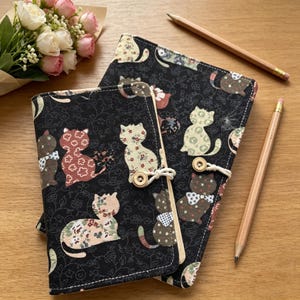 May include: Two black fabric journals with a cat pattern. The journals feature various cat designs in floral and polka dot patterns and are closed with wooden buttons and string. Two pencils and a bouquet of flowers are also present.