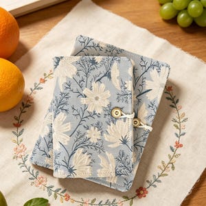 Puede incluir: Dos cuadernos rectangulares cubiertos de tela con un fondo azul claro y un patrón repetitivo de flores color crema y tallos azul oscuro. El cuaderno más pequeño está asegurado con un cordón elástico blanco y un pequeño botón redondo de color claro.
