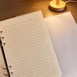 Puede incluir: Cuaderno abierto con p&aacute;ginas rayadas y una p&aacute;gina en blanco al lado. El cuaderno est&aacute; abierto en una p&aacute;gina con la palabra "DATE" visible. Una peque&ntilde;a l&aacute;mpara de madera con una pantalla plisada est&aacute; en el fondo.