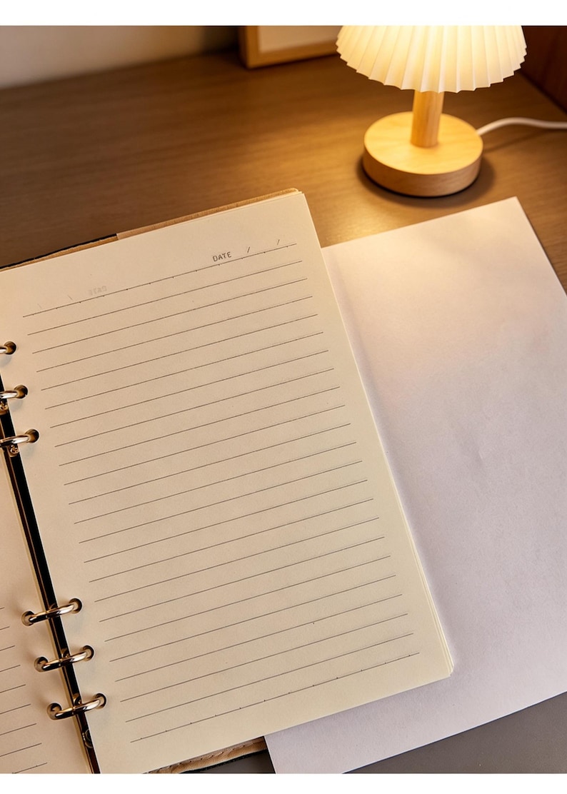 Puede incluir: Cuaderno abierto con p&aacute;ginas rayadas y una p&aacute;gina en blanco al lado. El cuaderno est&aacute; abierto en una p&aacute;gina con la palabra "DATE" visible. Una peque&ntilde;a l&aacute;mpara de madera con una pantalla plisada est&aacute; en el fondo.
