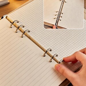 Puede incluir: Cuaderno abierto con papel rayado y cuadriculado. El cuaderno tiene una cubierta marr&oacute;n claro y anillos met&aacute;licos plateados. Se muestra una mano abriendo los anillos para agregar o quitar p&aacute;ginas. Las p&aacute;ginas son de color crema.