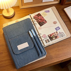 Puede incluir: Un planificador cubierto de mezclilla con un bolsillo y dos bol&iacute;grafos azul claro. El planificador est&aacute; abierto, revelando p&aacute;ginas rayadas con pegatinas decorativas y una impresi&oacute;n floral. El planificador est&aacute; sobre una superficie de madera.