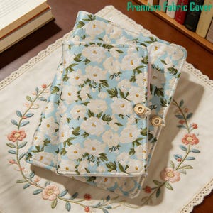 Puede incluir: Dos diarios cubiertos de tela con un estampado floral azul claro y cierres de botón de madera. Los diarios están apilados sobre un mantel individual bordado de color crema. El texto "Premium Fabric Cover" es visible en la esquina superior derecha.