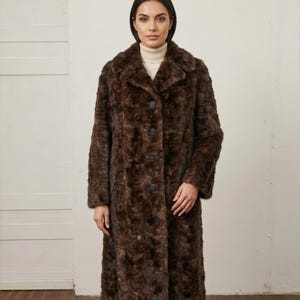May include: A long, brown faux fur coat with a classic collar and button closure. The coat has a textured appearance and a knee-length hem. The model is wearing a cream-colored turtleneck underneath the coat.