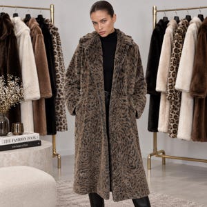 May include: A long, brown faux fur coat featuring a dark, swirling animal-inspired pattern, a wide collar, and long sleeves. The coat is worn over a black turtleneck and black pants, with other various coats hanging on racks in the background.