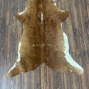 May include: A cowhide rug featuring a natural, irregular shape with a mottled pattern of warm brown and creamy white fur. The hide displays a dense, short-haired texture with a distinct color distribution across its surface, showcasing organic markings.