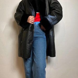 May include: A black leather coat with a black faux fur lining and cuffs, worn over a black mock neck jumper with a red and white horizontal stripe detail, paired with blue denim jeans. The coat features a button-front closure and long sleeves.