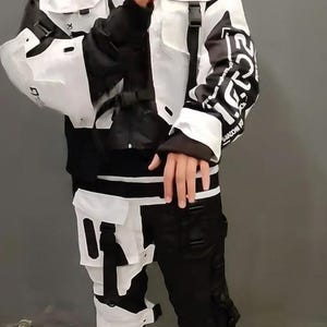 May include: A black and white jacket and pants set with multiple pockets and straps. The jacket has the letters "H-G" on the chest. The outfit is paired with black sneakers.