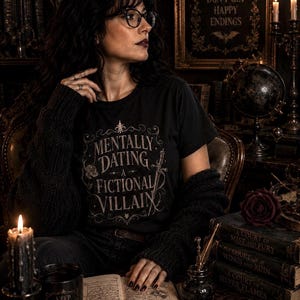 Puede incluir: Una persona con una camiseta negra con la frase "MENTALLY DATING A FICTIONAL VILLAIN" est&aacute; en una habitaci&oacute;n oscura. Se ven libros, velas y un letrero que dice "VILLAINS DON'T GET HAPPY ENDINGS".