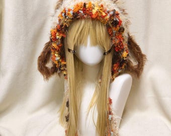 May include: A fuzzy, textured bonnet featuring a mix of cream, brown, orange, and yellow yarn. The hood has long, dangling braided ties and ear-like side accents, displayed on a mannequin head with long, straight blonde hair.