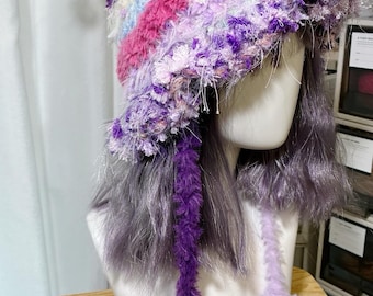 May include: A pointed crochet hat featuring a fuzzy, multi-colored texture with horizontal stripes in shades of purple, pink, blue, and white. The hat includes two long, dangling braided ties with star-shaped ends in dark purple and light lavender.