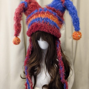 May include: A fuzzy, multi-colored jester-style hat featuring two long, pointed ear-like extensions topped with orange and multi-colored pom-poms. The hat is knit with red, blue, and orange yarn, and includes two long, braided tassels hanging down.