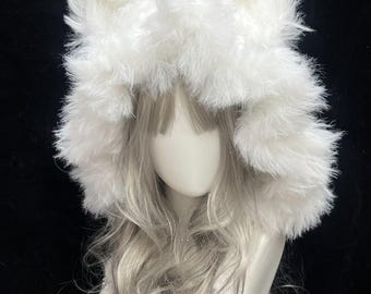 May include: A fluffy white faux fur hood with large, upright ears. The hood is designed to resemble a cat or fox. The hood is worn over a wig of long, wavy, silver hair. The background is black.