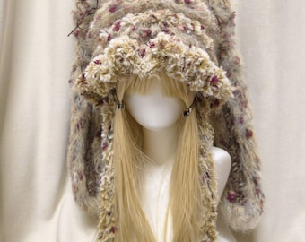 May include: A fuzzy, textured hat with long, drooping ear-like flaps, crafted from a variegated yarn in shades of cream, tan, and magenta. The hat features a ruffled brim and two long, braided ties hanging down from the sides of the head.