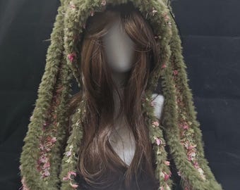 May include: A fuzzy olive green hat with long, dangling ear-like flaps featuring pink and cream textured yarn accents. The hat includes two white pom-poms attached to the ends of thin, matching green cords hanging from the front of the headpiece.