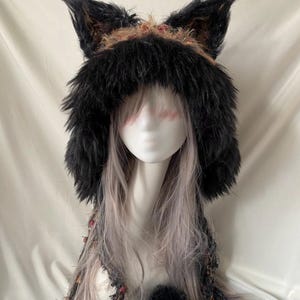 May include: A black faux fur hood featuring prominent animal-like ears on top. The hood is lined with a textured, multi-colored fabric containing shades of brown, orange, and black. Two long, dangling fabric ties with black pom-poms hang down.