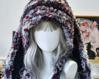 May include: A fluffy, hooded hat with long ear flaps. The hat is primarily black, white, and gray, with small red accents. The hat is made of a soft, textured material, perfect for cold weather. The hat is displayed on a mannequin head.