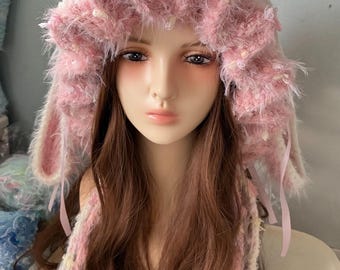 May include: A fuzzy, textured bonnet in shades of pink and white, featuring long, dangling ear-like side panels. The hat is crafted from a soft, shaggy yarn material and includes thin, light pink ribbon ties hanging down from the sides.