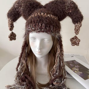 May include: A brown and white crocheted jester hat with three points, each ending in a star-shaped embellishment. The hat is displayed on a mannequin head with long, brown and white hair. The hat is a unique, handmade accessory.