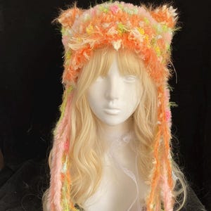 May include: A handmade, fluffy cat ear hat in shades of orange, pink, and yellow. The hat features long, colourful strands that hang down. The hat is displayed on a mannequin head with blonde hair.